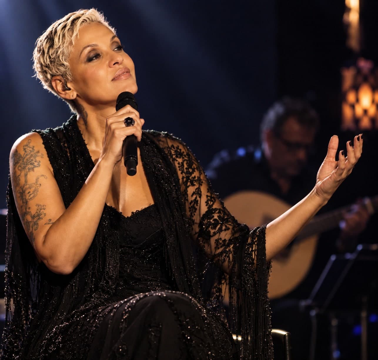 Mariza cantando ao vivo, sentada, microfone na mão e músicos ao fundo